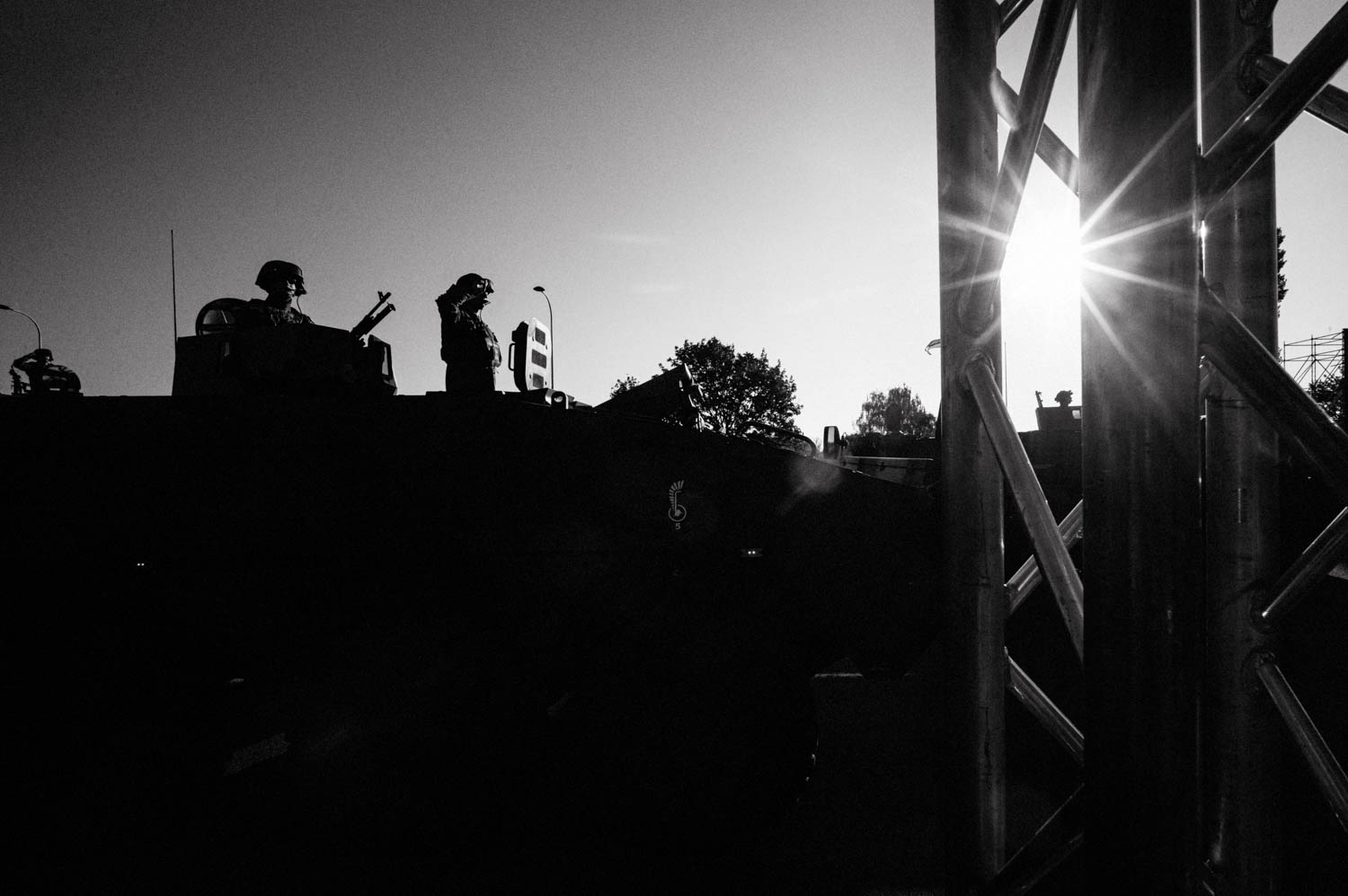 Military vehicles pass by during an early morning rehearsal for Poland’s Armed Forces Day parade. Soldiers salute on top. Sun glints through metal.